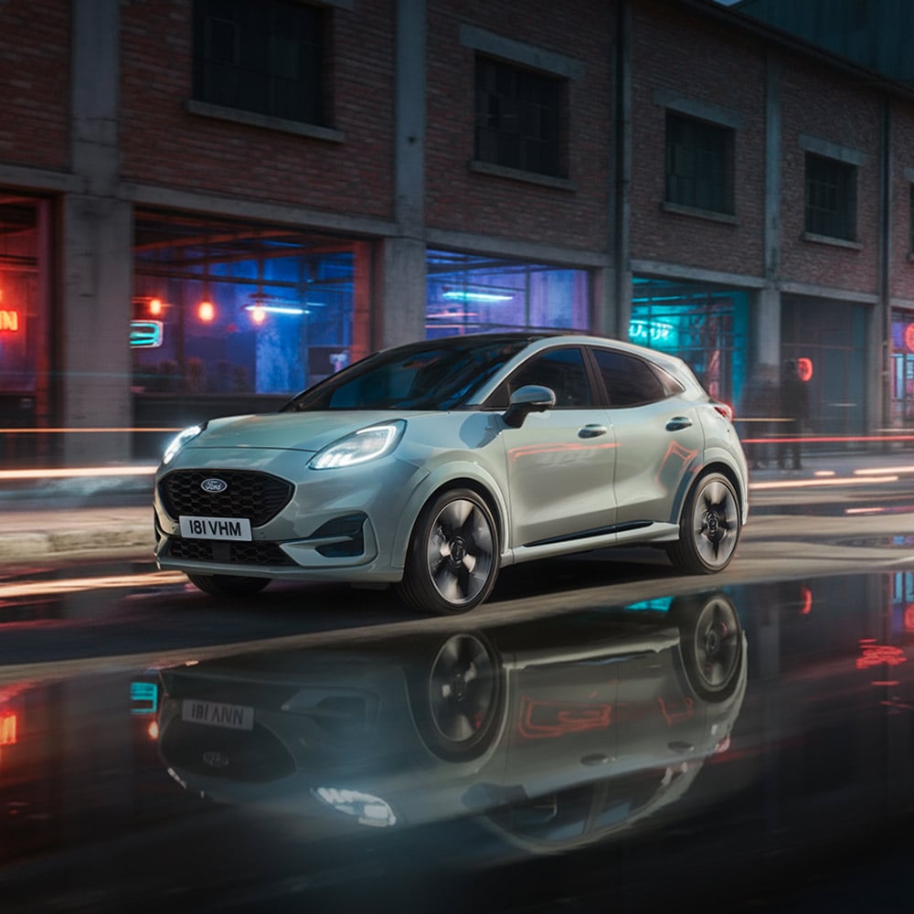 Ανοιχτόχρωμο Ford Puma οδηγεί σε υγρό αστικό δρόμο τη νύχτα, με αντανακλάσεις νέον.