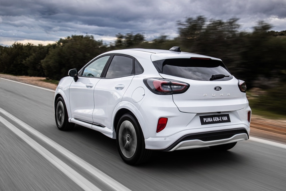 Πίσω τριών τετάρτων άποψη λευκού Ford Puma Gen-E Van που κινείται σε δρόμο, με θολωμένο φυσικό φόντο.