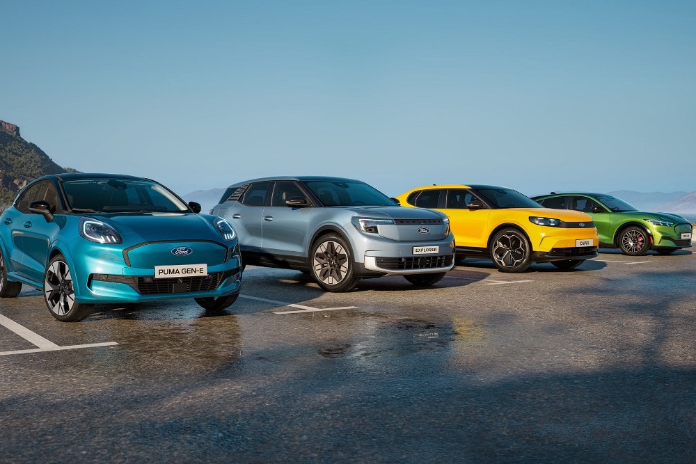 Τέσσερα ηλεκτρικά μοντέλα της Ford σε διάφορα χρώματα (γαλάζιο Puma Gen-E, ασημί Explorer, κίτρινο Capri και πράσινο Mustang Mach-E) παρκαρισμένα δίπλα-δίπλα με φόντο βουνά