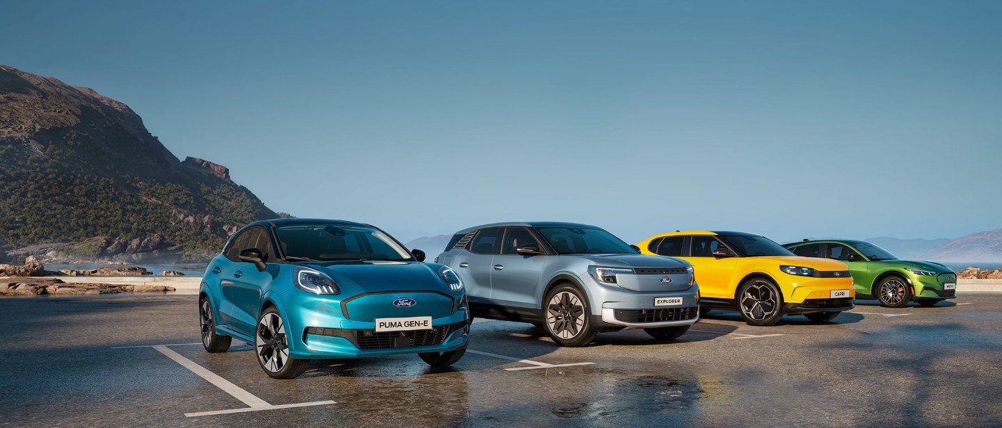 Τέσσερα ηλεκτρικά μοντέλα της Ford σε διάφορα χρώματα (γαλάζιο Puma Gen-E, ασημί Explorer, κίτρινο Capri και πράσινο Mustang Mach-E) παρκαρισμένα δίπλα-δίπλα με φόντο βουνά