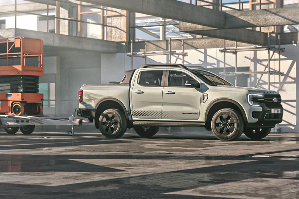 Ένα γκρι Ford Ranger σε εργοτάξιο. Ρυμουλκεί πλατφόρμα μεταφοράς που φέρει ανυψωτικό τύπου scissor lift. Στο υπόβαθρο φαίνονται σκυρόδεμα, μεταλλικές δοκοί, σκαλωσιές και κολώνες εργοταξίου, με φωτεινό ήλιο και σκιές.
