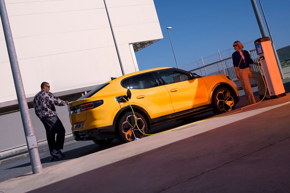 Άτομα φορτίζουν ένα κίτρινο Ford Capri EV σε δημόσιο σταθμό φόρτισης. 