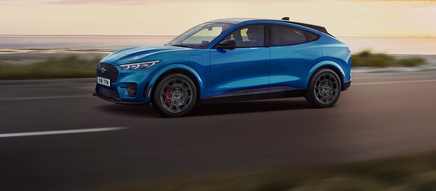 Ένα μπλε ηλεκτρικό Ford Mustang Mach-E καθώς κινείται κατά μήκος ενός παραλιακού δρόμου το ηλιοβασίλεμα.