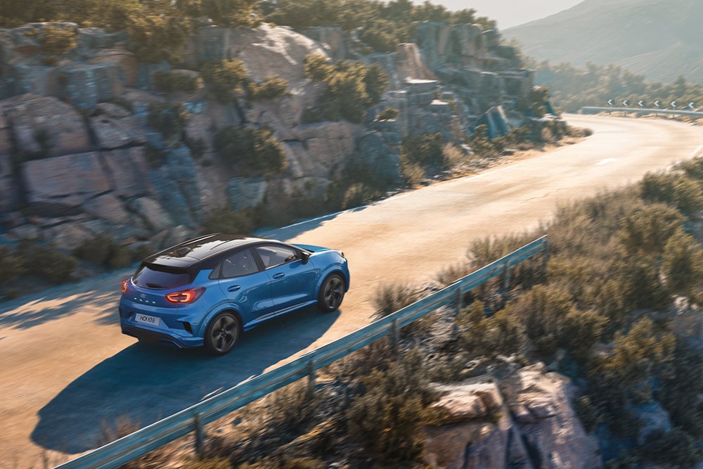 Ένα μπλε Puma Gen-E που κινείται κατά μήκος ενός ελικοειδούς ορεινού δρόμου.