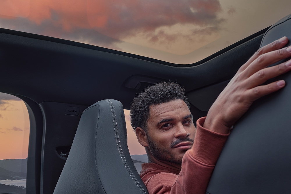 Ένας άνδρας καθισμένος μέσα σε αυτοκίνητο στο ηλιοβασίλεμα, με το χέρι του να ακουμπά στην πλάτη ενός από τα καθίσματα.