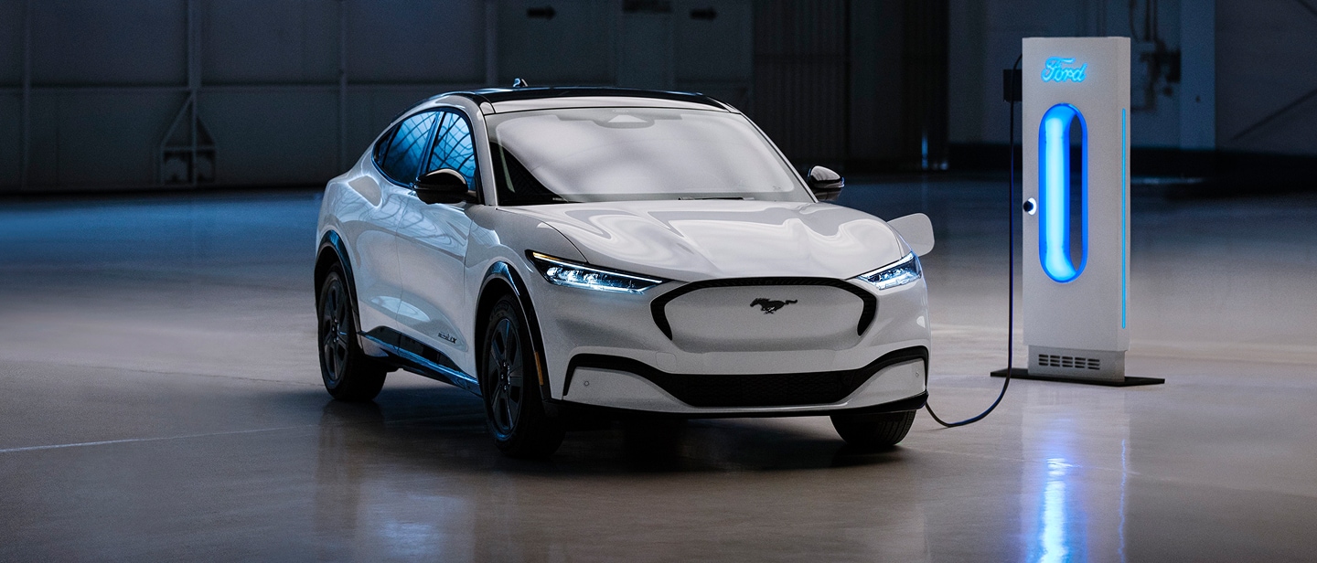 Ένα λευκό Ford Mustang Mach-E σταθμευμένο σε γκαράζ, συνδεδεμένο σε έναν λαμπερό μπλε σταθμό φόρτισης.