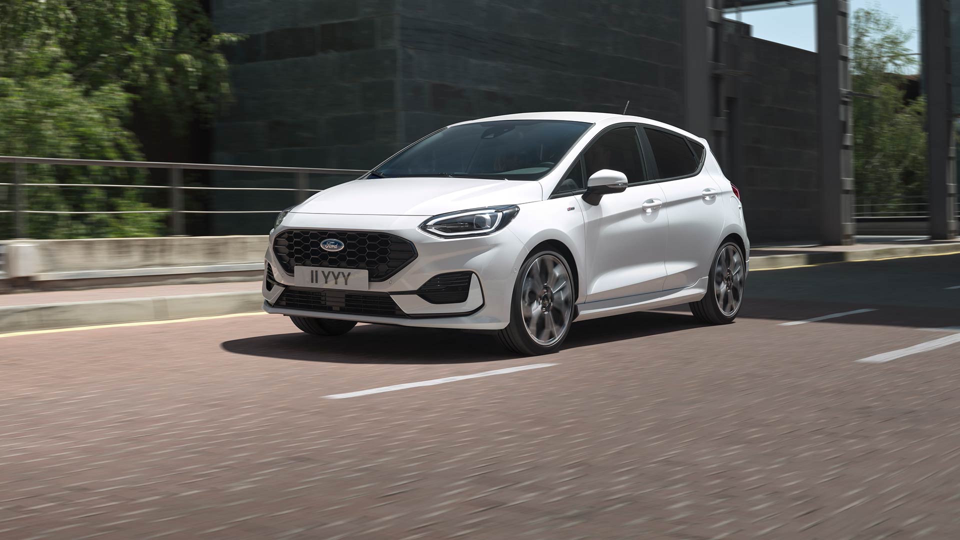 Λευκό Ford Fiesta που οδηγείται σε δρόμο πόλης, με πράσινο και κτίρια στο βάθος.