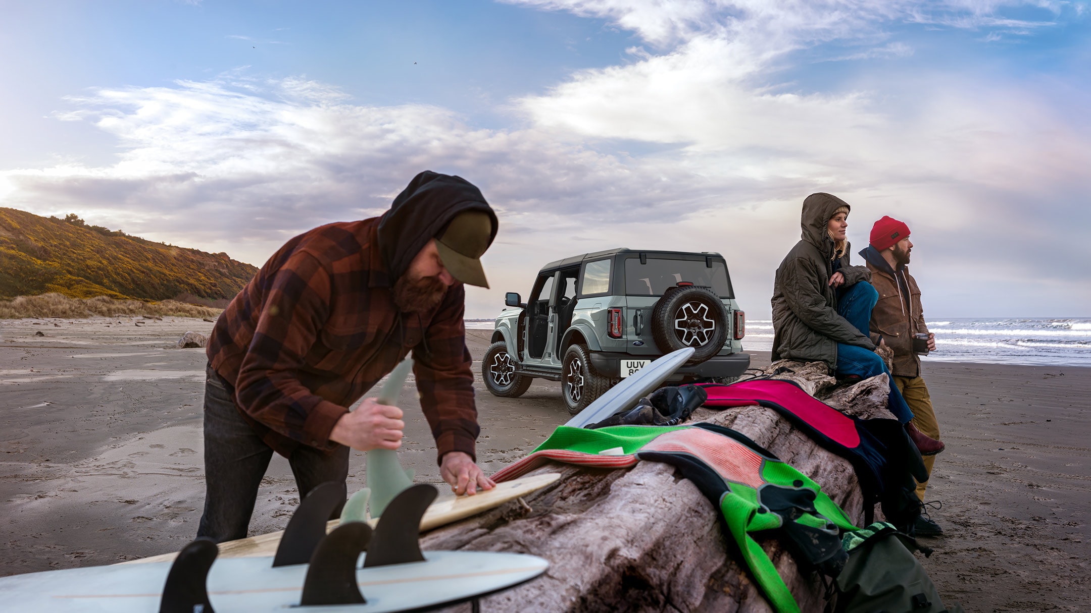 Ford Bronco on beach with three people sitting with surfboards