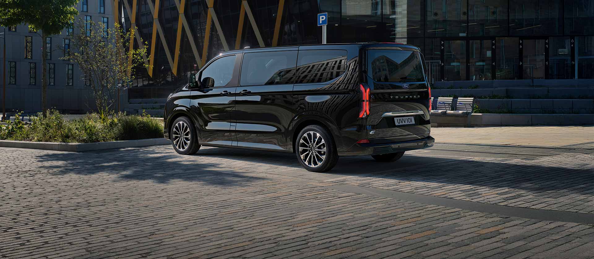 Μαύρο Ford E-Tourneo Custom σταθμευμένο μπροστά από ένα μοντέρνο κτήριο ενώ φαίνεται η πλαϊνή και πίσω πλευρά του.