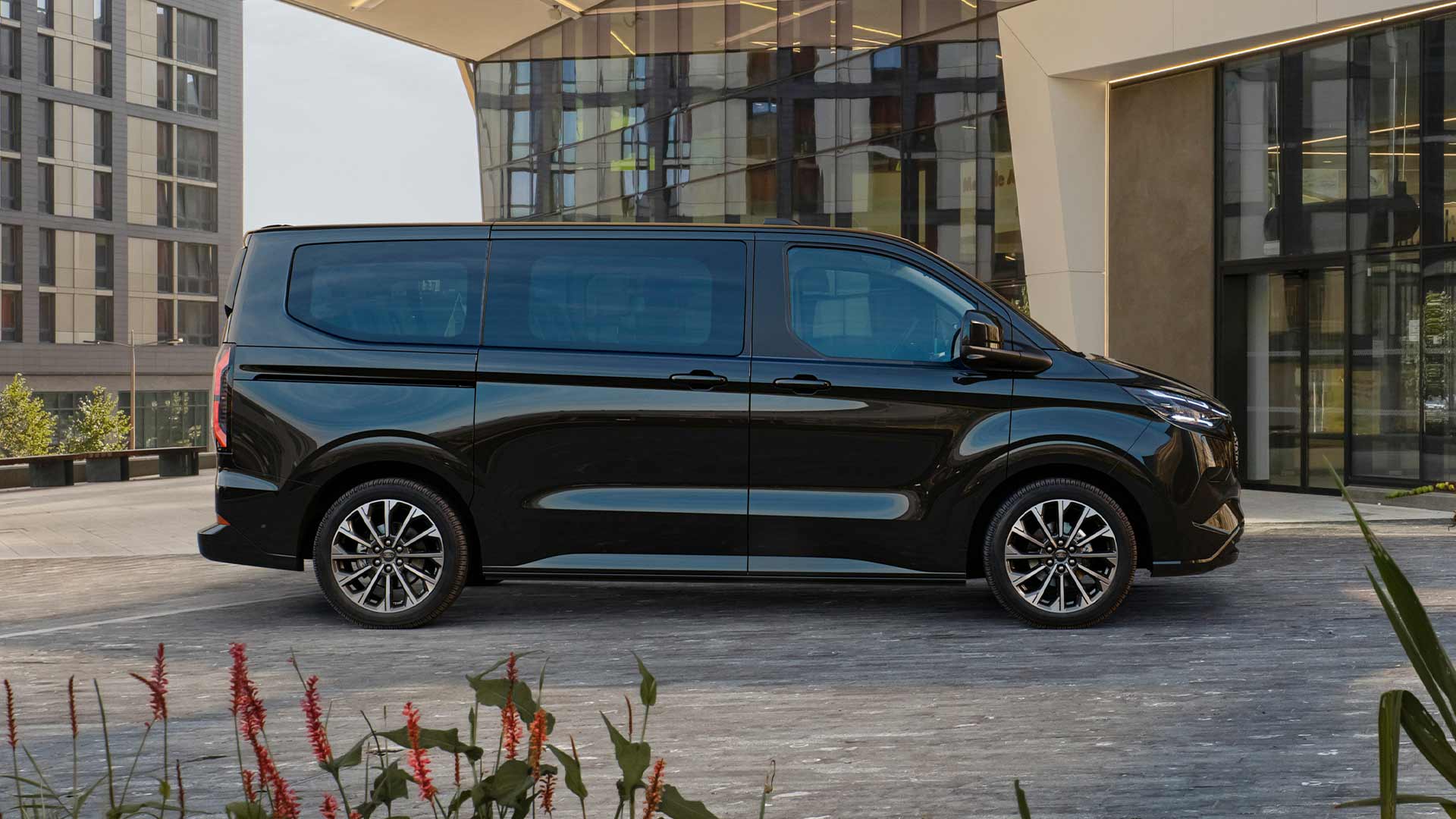 Μαύρο Ford E-Tourneo Custom σταθμευμένο μπροστά από ένα μοντέρνο κτήριο.
