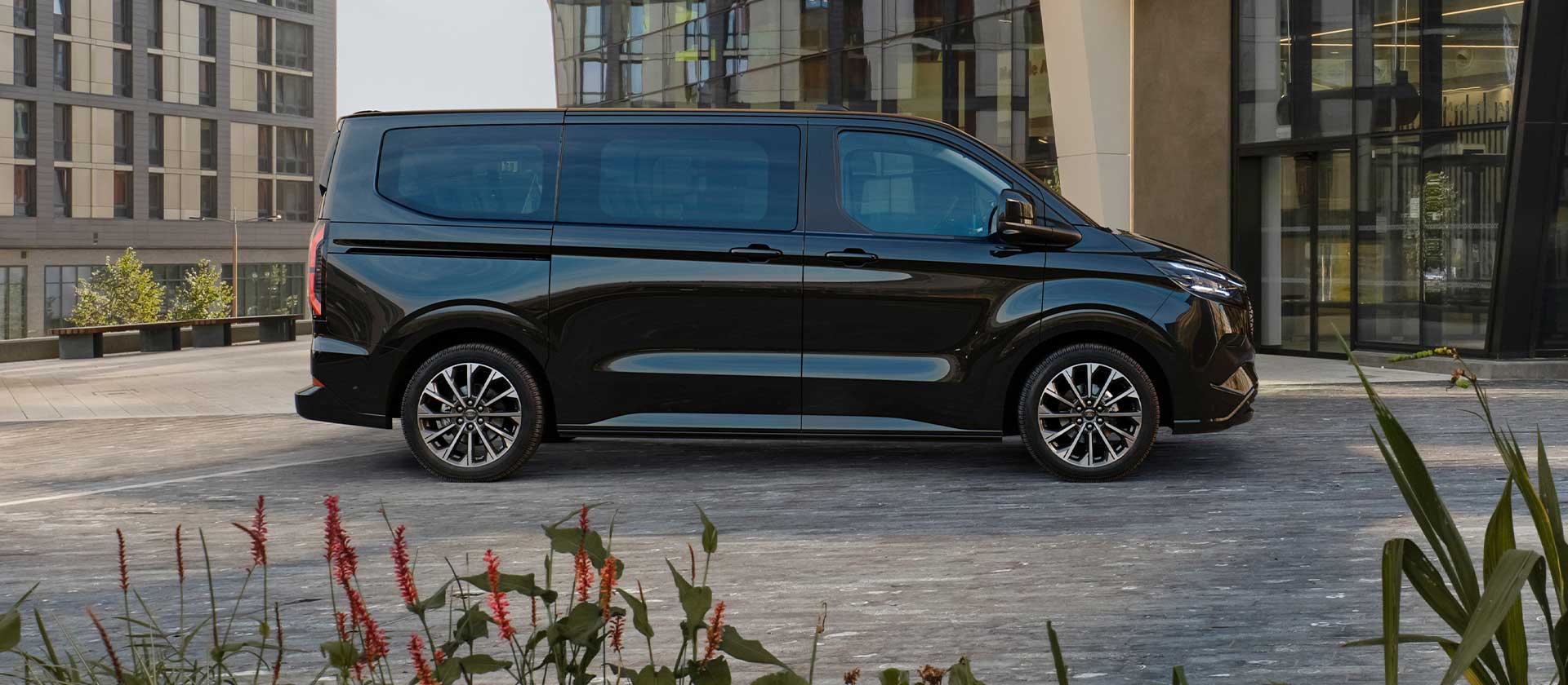 Μαύρο Ford E-Tourneo Custom σταθμευμένο μπροστά από ένα μοντέρνο κτήριο.