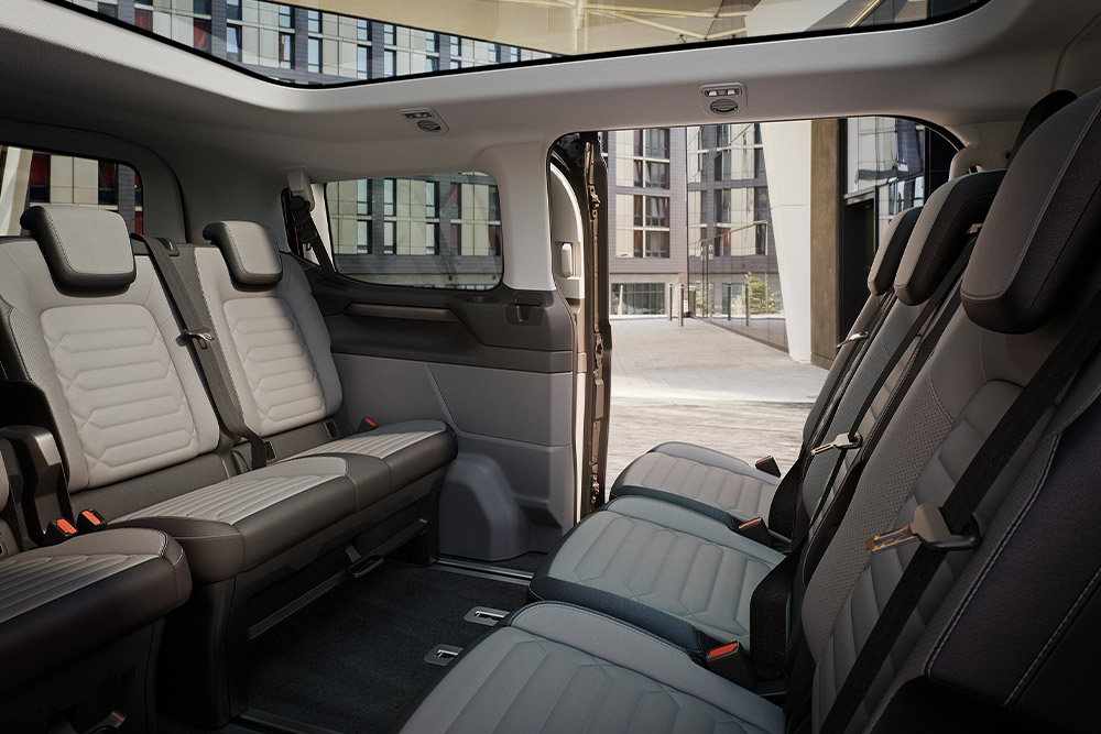 Εσωτερική άποψη του Ford Tourneo Custom, όπου φαίνονται πολλές σειρές γκρι καθισμάτων για τους επιβάτες, καθώς και η πανοραμική ηλιοροφή.