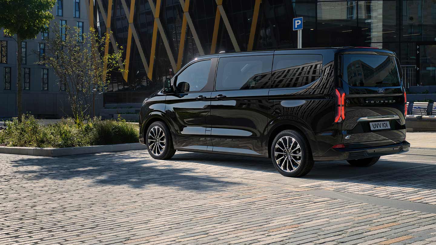 Μαύρο Ford E-Tourneo Custom σταθμευμένο μπροστά από ένα μοντέρνο κτήριο ενώ φαίνεται η πλαϊνή και πίσω πλευρά του.