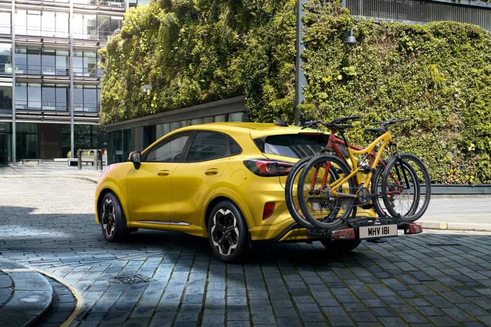 Ford Puma Gen-E σε χρώμα Electric Yellow παρκαρισμένο σε αστικό περιβάλλον με δύο ποδήλατα τοποθετημένα σε πίσω σχάρα ποδηλάτων.