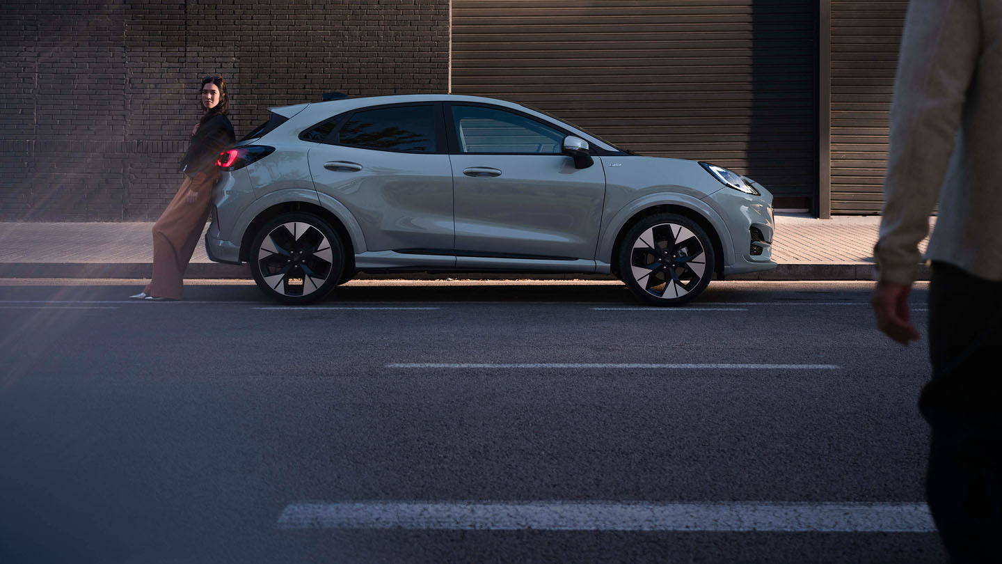 Side view of a light grey Ford Puma parked on a street, with a person leaning against the rear.