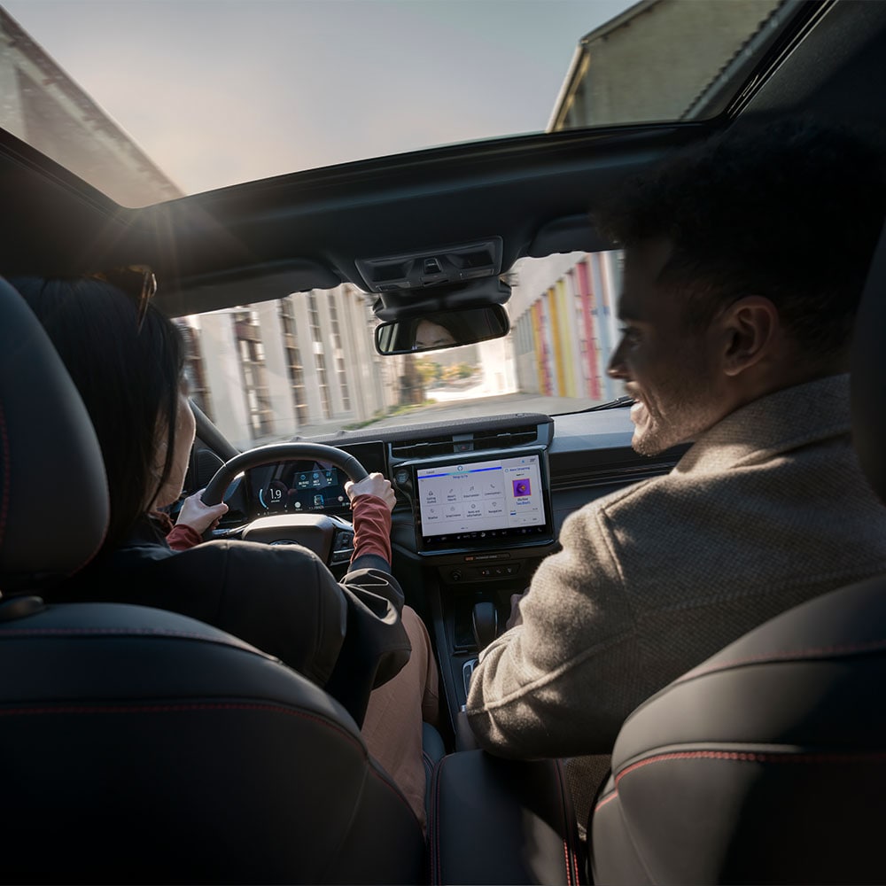 Ο οδηγός και ο επιβάτης συνομιλούν ενώ κινούνται σε ένα ηλιόλουστο τοπίο με κατοικίες.