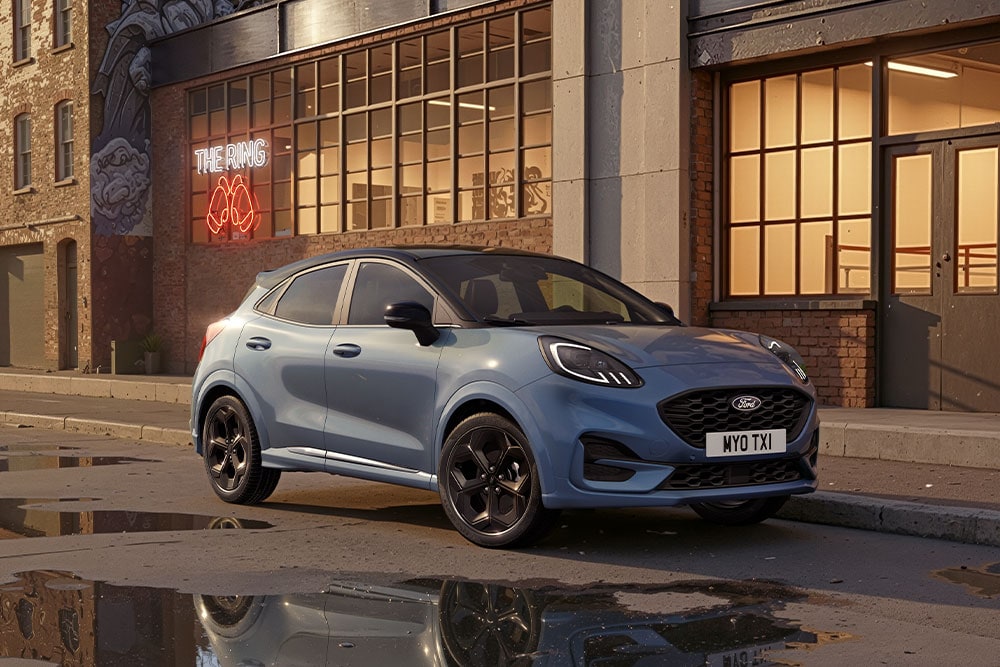 A blue Ford Puma parked on an urban street at sunset, with puddles reflecting the buildings.