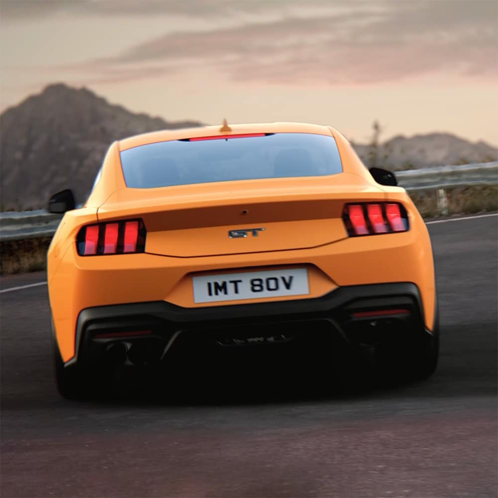 Το πίσω μέρος μιας Ford Mustang GT Fastback σε χρώμα Orange Fury που στρίβει σε μια κλειστή στροφή σε επαρχιακό δρόμο