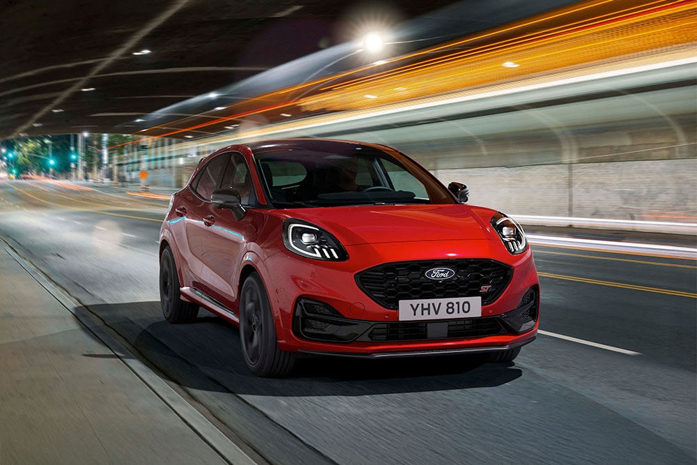 Ένα κόκκινο Ford Puma ST σπορ SUV που οδηγεί σε έναν έντονα φωτισμένο δρόμο της πόλης τη νύχτα.