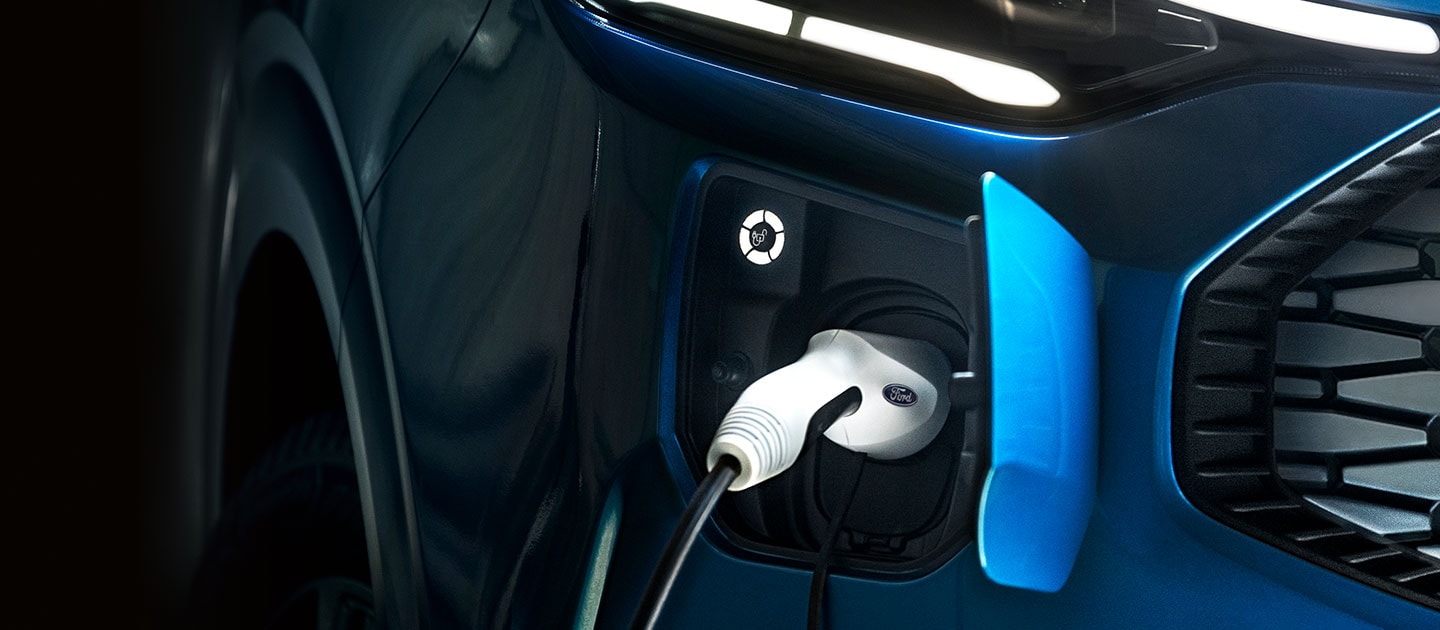 Κοντινή άποψη της θύρας φόρτισης ενός μπλε Ford Tourneo Custom, με το καλώδιο φόρτισης στην πρίζα.