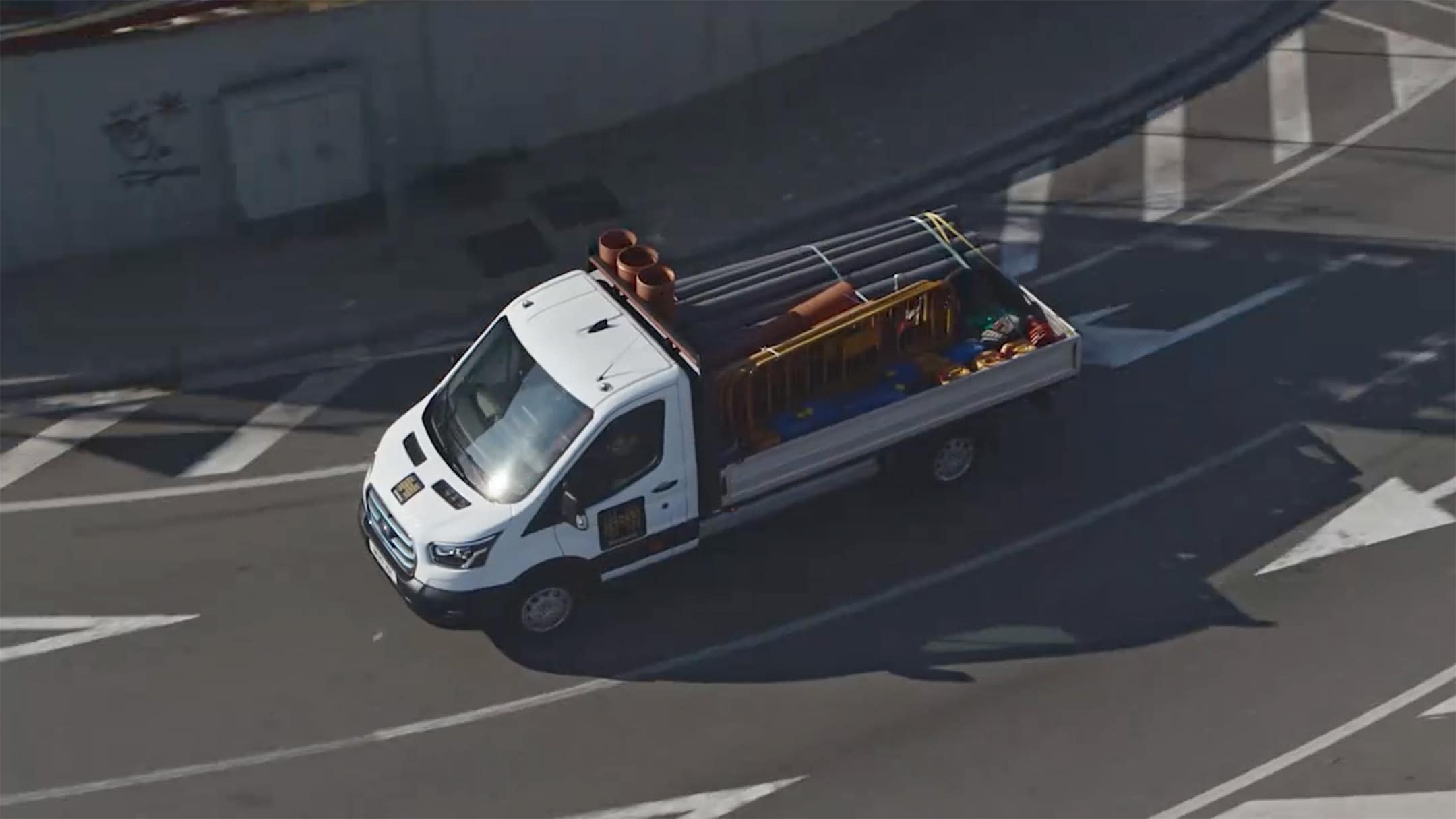 Πανοραμική λήψη Ford E-Transit Chassis που μεταφέρει οικοδομικά υλικά σε ασφαλτοστρωμένο δρόμο.