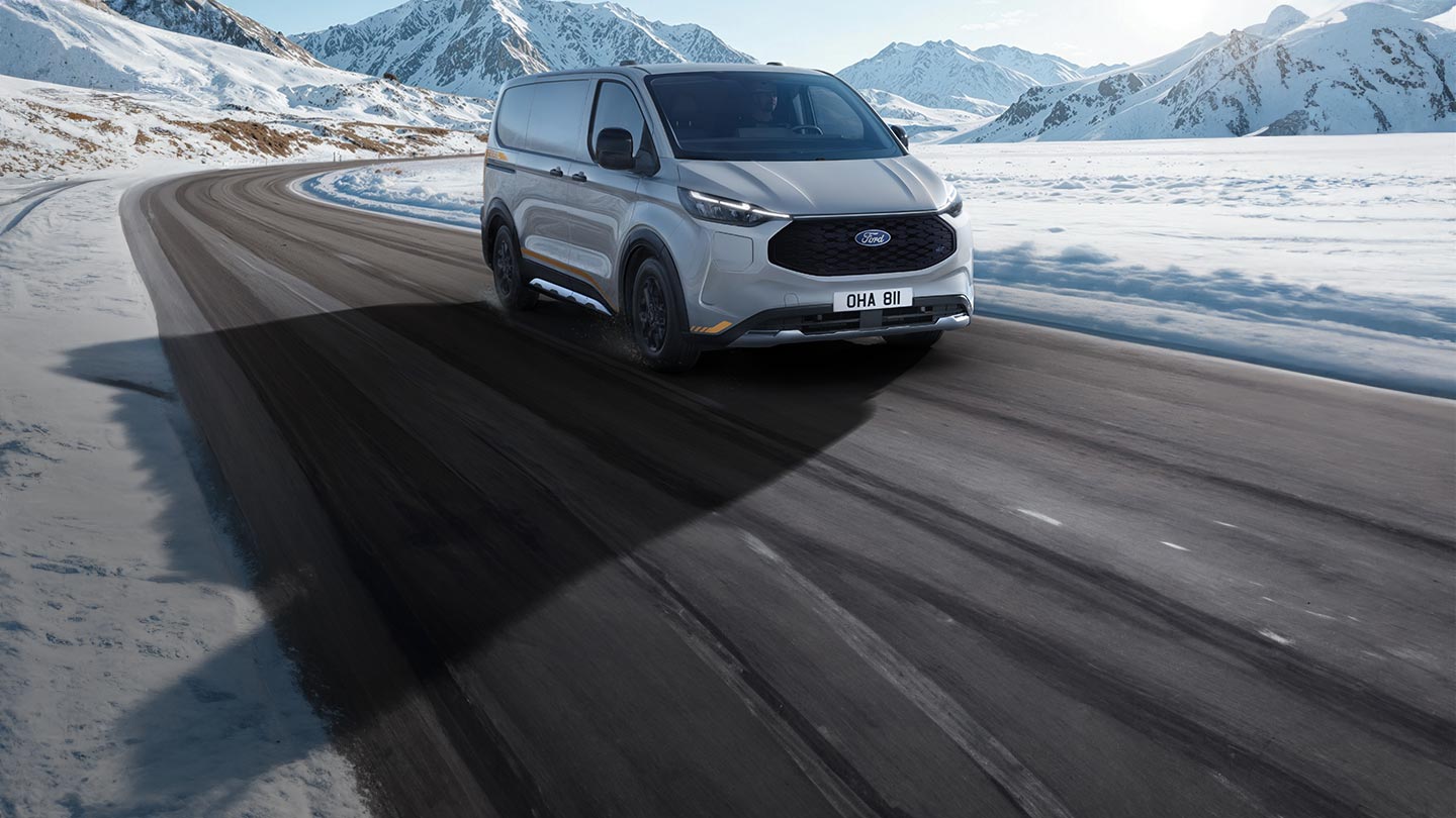  Ένα ανοιχτό γκρι Ford E-Transit Custom AWD βαν με πορτοκαλί λεπτομέρειες φαίνεται να οδηγεί σε έναν ελικοειδή δρόμο μέσα σε ένα χιονισμένο ορεινό τοπίο.