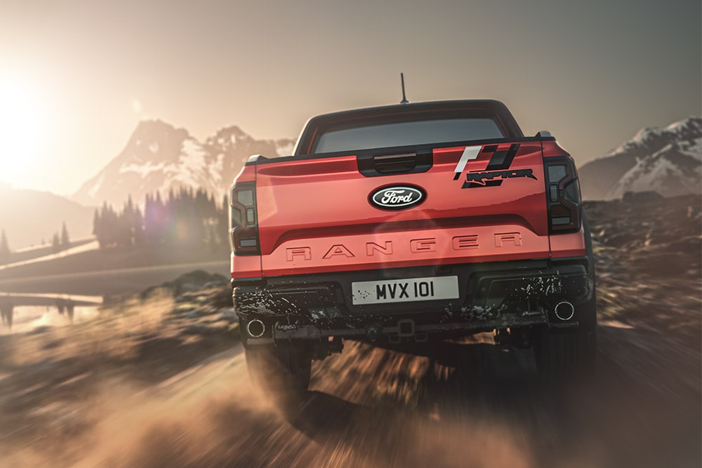 Πίσω όψη κόκκινου Ford Ranger Raptor που κινείται σε χωματόδρομο, με βουνά στο βάθος.