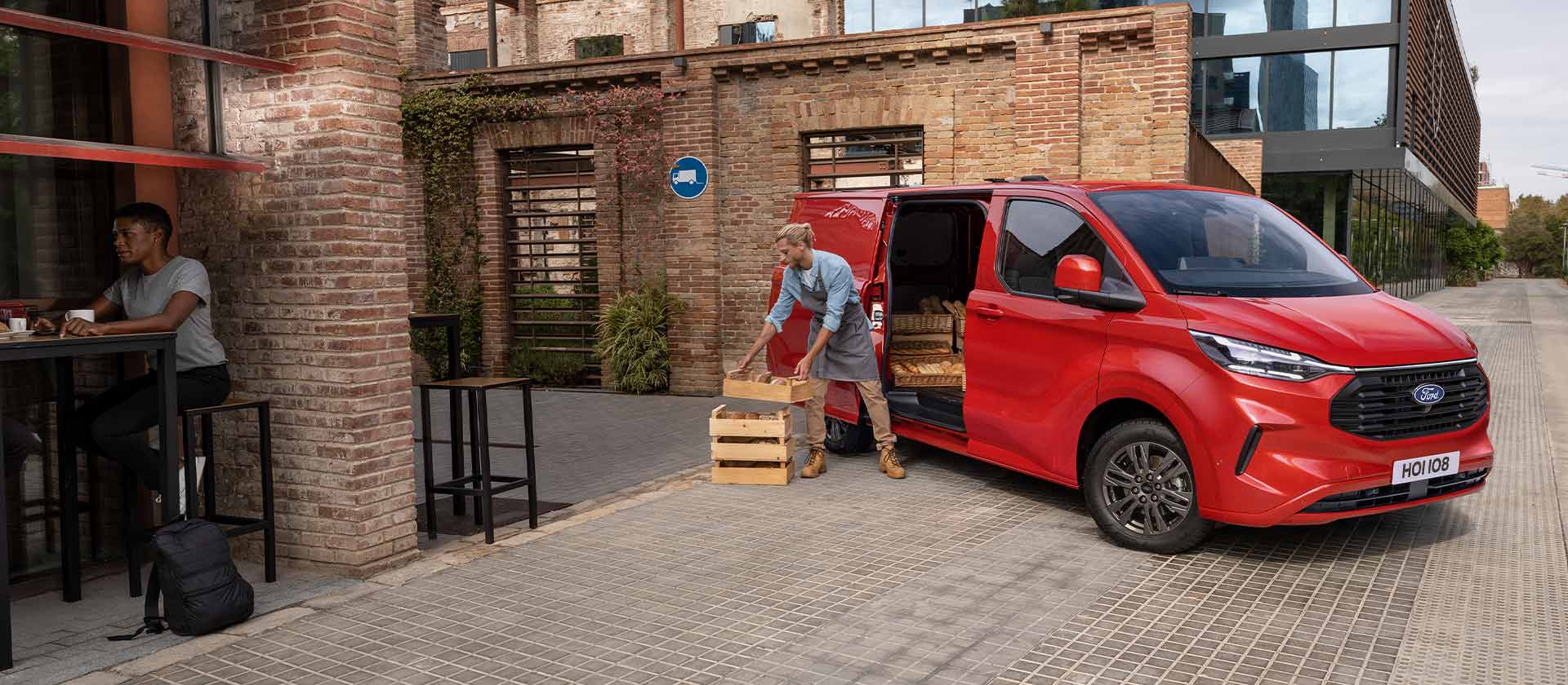 Άτομο ξεφορτώνει κιβώτια από ένα κόκκινο Ford Transit Custom van σταθμευμένο μπροστά από μια καφετέρια. 