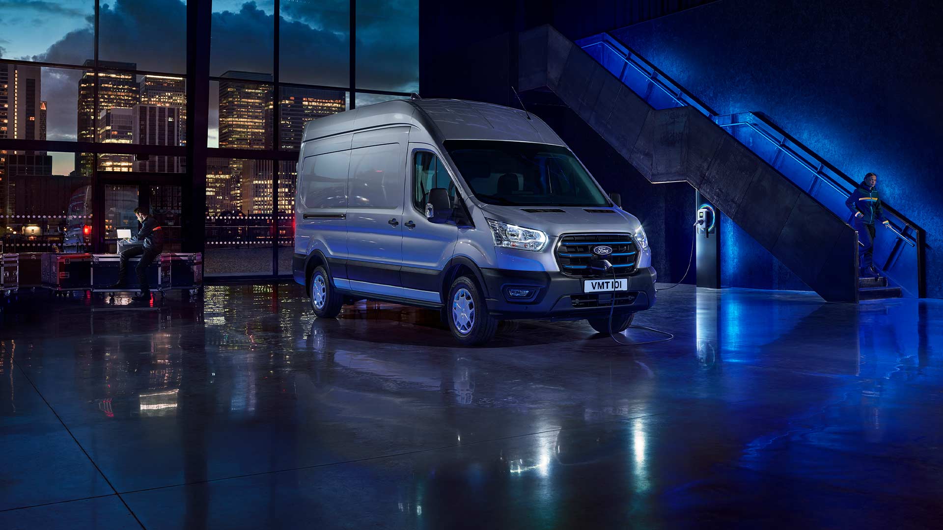 Ασημί Ford E-Transit ηλεκτρικό van σταθμευμένο σε εσωτερικό χώρο με ορατό τον ορίζοντα της πόλης μέσα από μεγάλα παράθυρα.