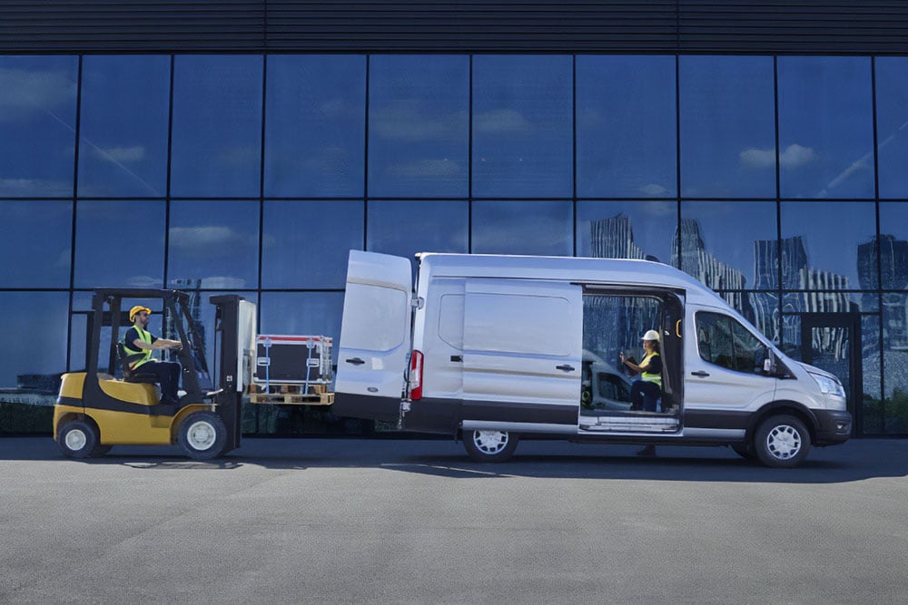 Δύο άτομα που φορτώνουν φορτίο σε ένα ασημί Ford Transit van με περονοφόρο ανυψωτικό όχημα.