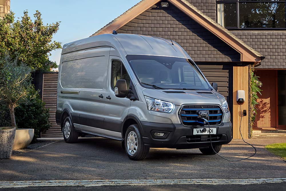 Ασημί Ford  E-Transit van σταθμευμένο σε ιδιωτικό δρόμο δίπλα σε ένα σπίτι, συνδεδεμένο με έναν σταθμό φόρτισης.