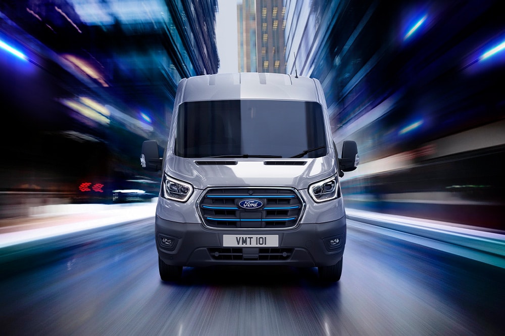 Μπροστινή όψη ενός γκρι βαν Ford Transit με μπλε λεπτομέρειες στη μάσκα, το οποίο κινείται με ταχύτητα μέσα στην πόλη τη νύχτα.
