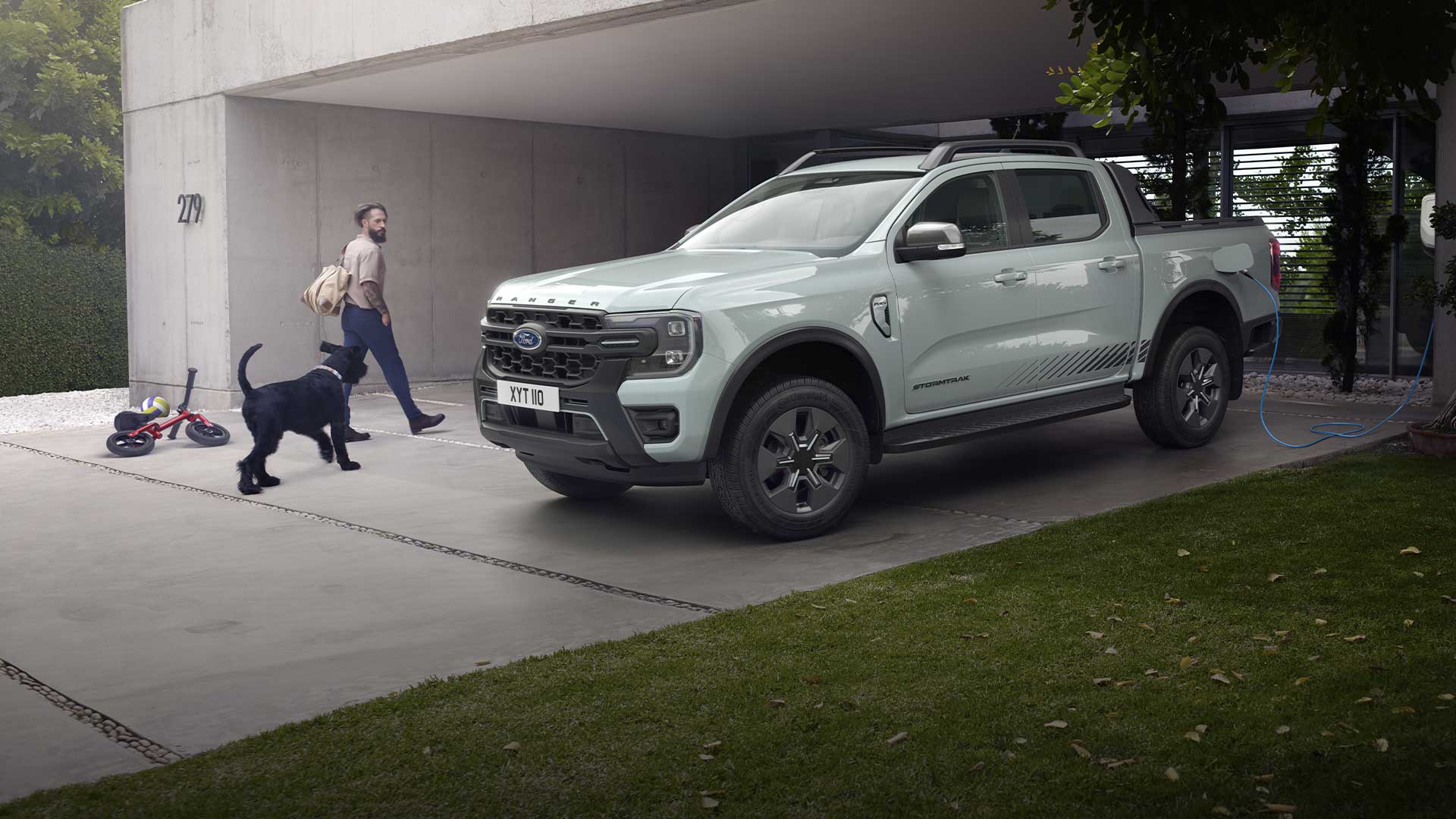 Ένας άνδρας και ένας σκύλος περπατούν προς ένα γκρι pick-up φορτηγό Ford Ranger Plug-in υβριδικό που φορτίζει σε έναν δρόμο. 