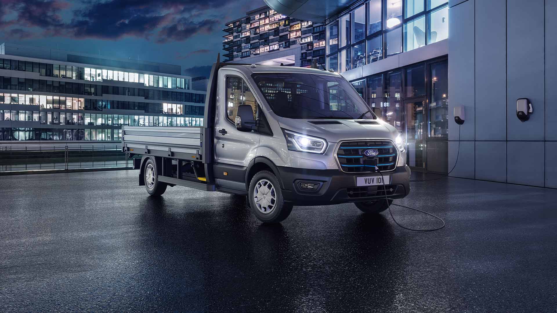 Ασημί Ford Transit Chassis Cab σταθμευμένο μπροστά από ένα μοντέρνο κτίριο τη νύχτα.