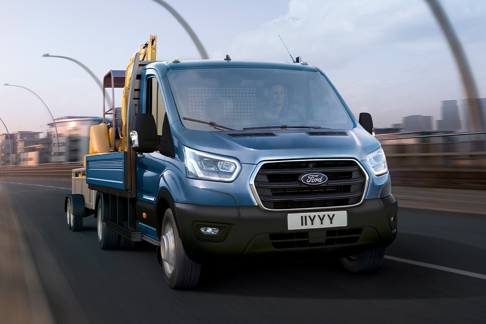 Άποψη της μπροστινής μάσκας ενός Ford Transit Chassis Cab που μεταφέρει έναν εκσκαφέα και κινείται σε έναν δρόμο πόλης. 