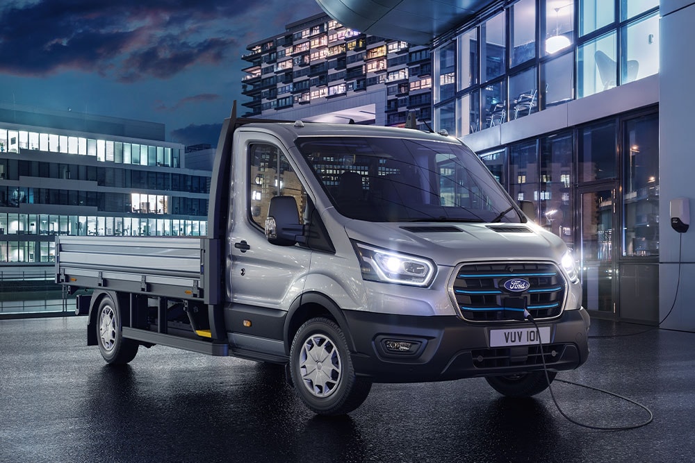 Ασημί Ford E-Transit Chassis Cab σταθμευμένο μπροστά από ένα μοντέρνο κτίριο τη νύχτα.