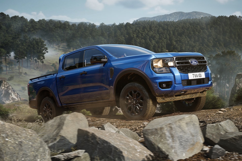 Μπλε φορτηγό Ford Ranger κινείται κατά μήκος ενός χωματόδρομου μέσα σε ένα δάσος, αφήνοντας ίχνη σκόνης.