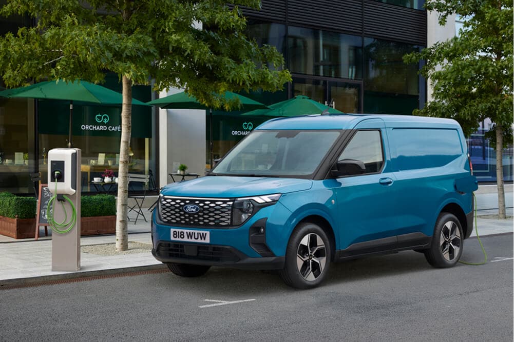 Ένα μπλε Ford E-Transit Courier από την μπροστινή τρία τέταρτα γωνία, παρκαρισμένο δίπλα σε σταθμό φόρτισης.