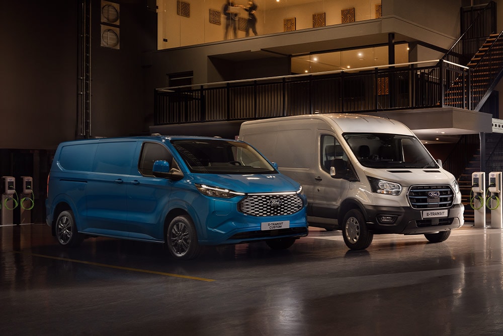 Ένα μπλε Ford E-Transit Custom και ένα ασημί Ford E-Transit φορτίζουν σε χώρο στάθμευσης αυτοκινήτων.