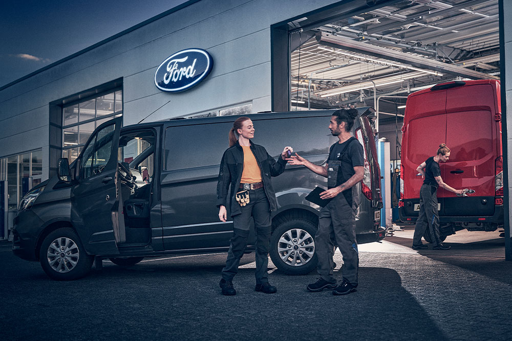 Δύο εργαζόμενοι της Ford παραδίδουν κλειδιά έξω από συνεργείο, με ένα κόκκινο βαν στο βάθος, το σούρουπο.